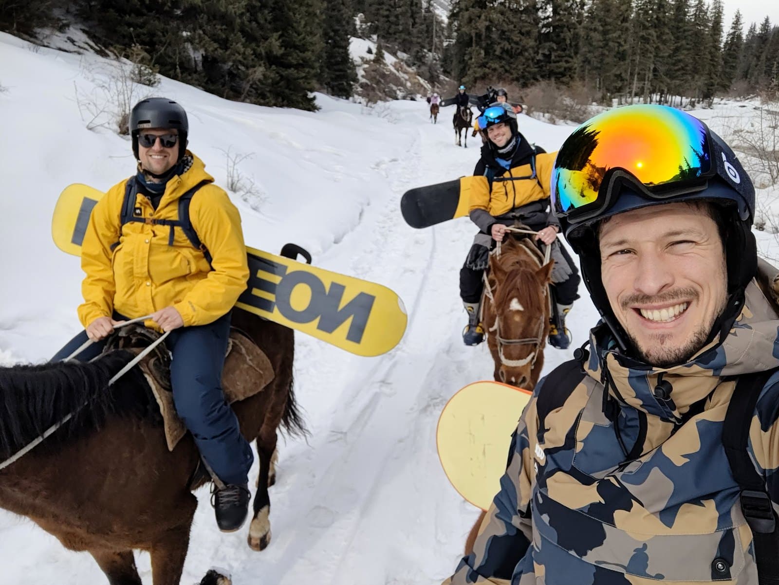 Three of the group riding through deep snow with snowboards strapped across their backs, grinning at the camera.