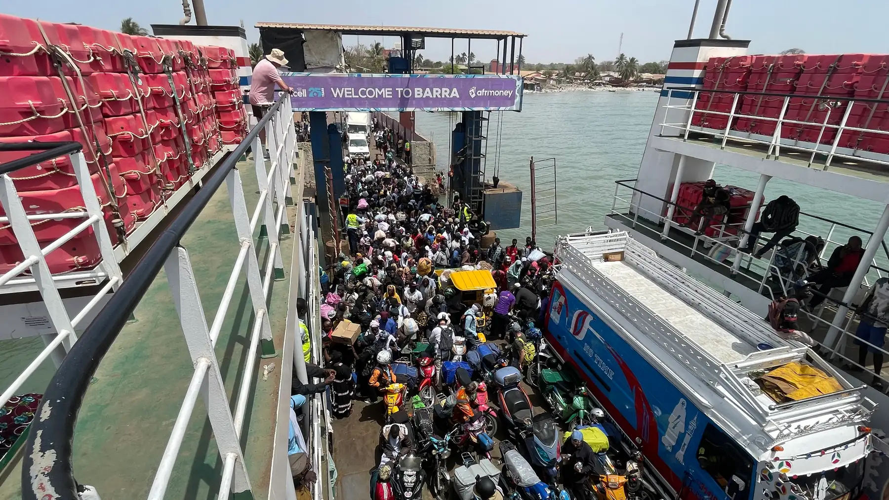 Looking down from the upper deck of a ferry onto a crowd of foot passengers, loaded motorbikes and a painted bus, with a banner reading WELCOME TO BARRA over the ramp.