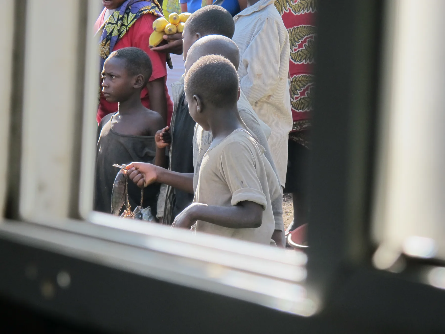A child holds up a small fish by the tail, seen through a window with several people behind them.