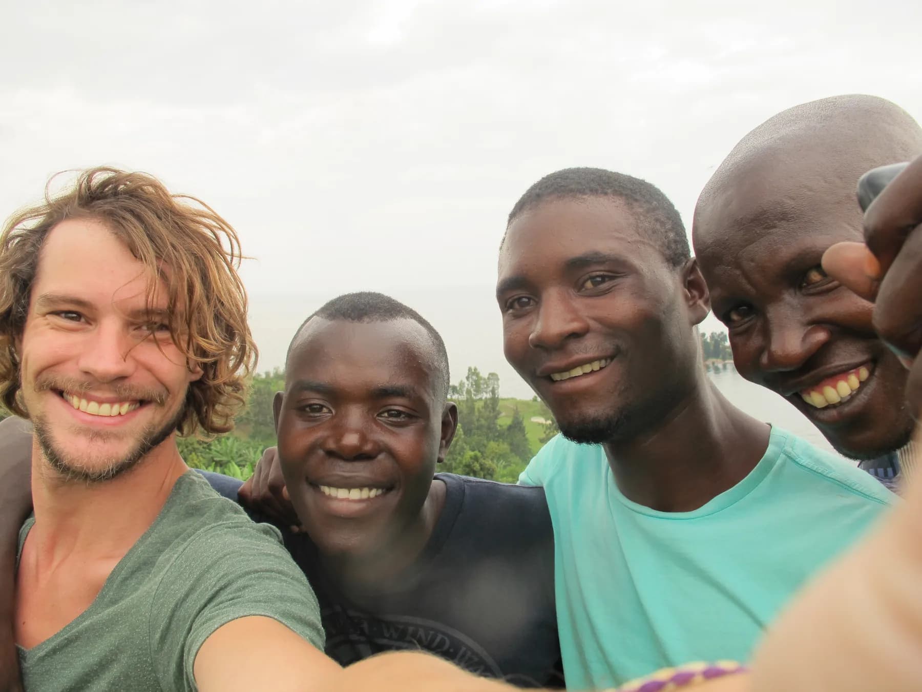 Four men lean together for a selfie, smiling, with greenery and hills behind them under an overcast sky.