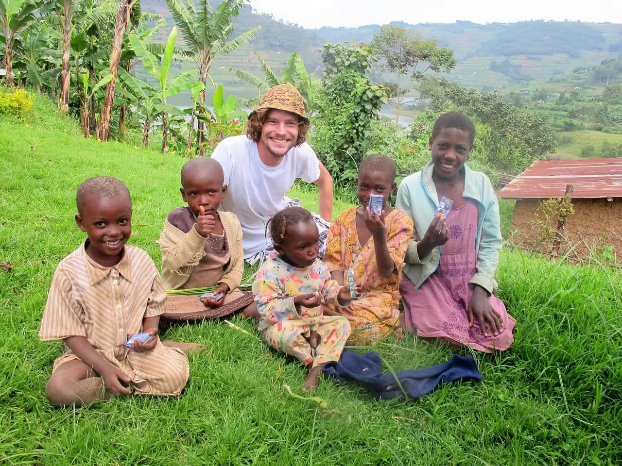 A man crouches behind five children sitting on a grassy slope, with banana plants and a corrugated roof behind them.