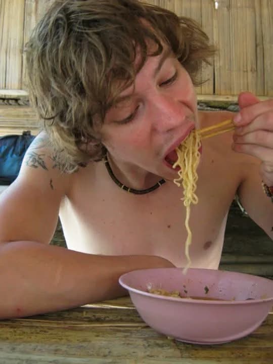 A shirtless young man slurps a long mouthful of noodles from a pair of chopsticks over a pink plastic bowl at a wooden table.