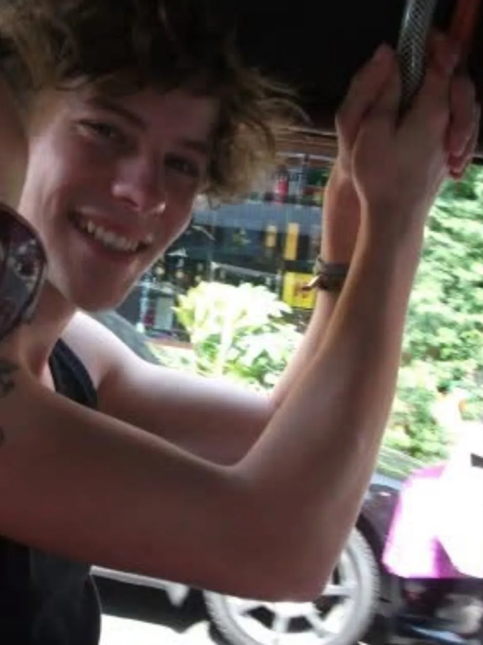 A young man grins over his shoulder in an open-sided ride, one arm up holding the rail overhead, with a car wheel and shopfronts blurring past behind him.