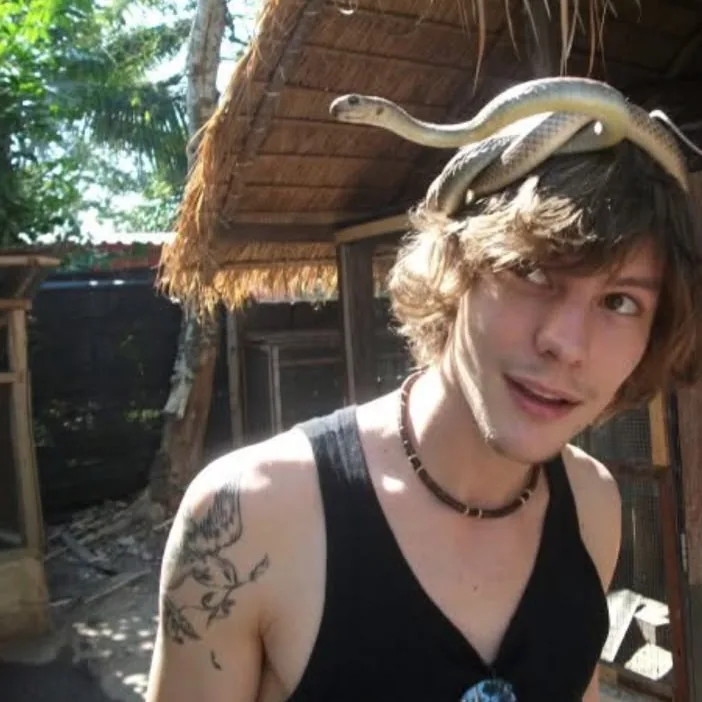 A young man in a black vest smiles at the camera with a pale snake draped across the top of his head, beneath the thatched eaves of a wooden hut.