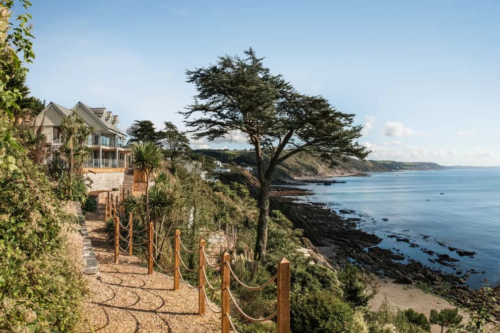 A coastal house and garden path above a beach and blue sea in Cornwall.